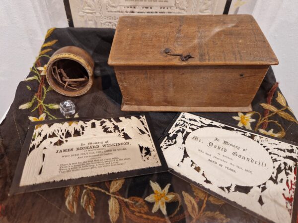 Small rectangular wooden box next to a cylindrical pot containing twigs. In front of the box are two funeral cards, white with black edging and details of funeral. 