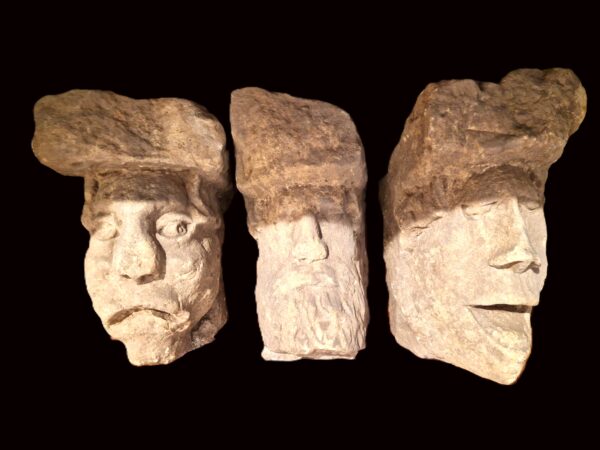 Three stone heads side by side, the left one has adownturned mouth and round nose, central one has a long face with poorly defined features, the face on the right has an open mouth and pointed chin. All male heads. 