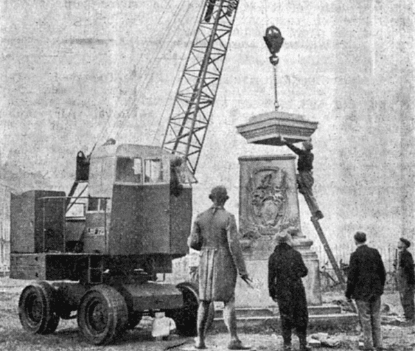 a small crane on wheels lifting a square cap onto the top of a statue plinth. Next to the plinth stands a larger than lifesize statue of Captain Cook. a man disobeying all modern health and safety ladder regulations is standing on a ladder moving the top into place as he is watched by 3 other workmen. This is the erection of the Captain Cook Monument on Whitby West Cliff. 